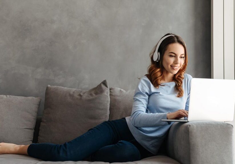 Portrait of a cheerful young woman listening to music with headphones and using laptop computer while relaxing on a couch at home